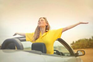 A young woman enjoying the wind as she drives her convertible car with her arms outstretched.