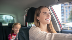 A smiling woman driving a car with a young girl in the backseat, both looking happy.
